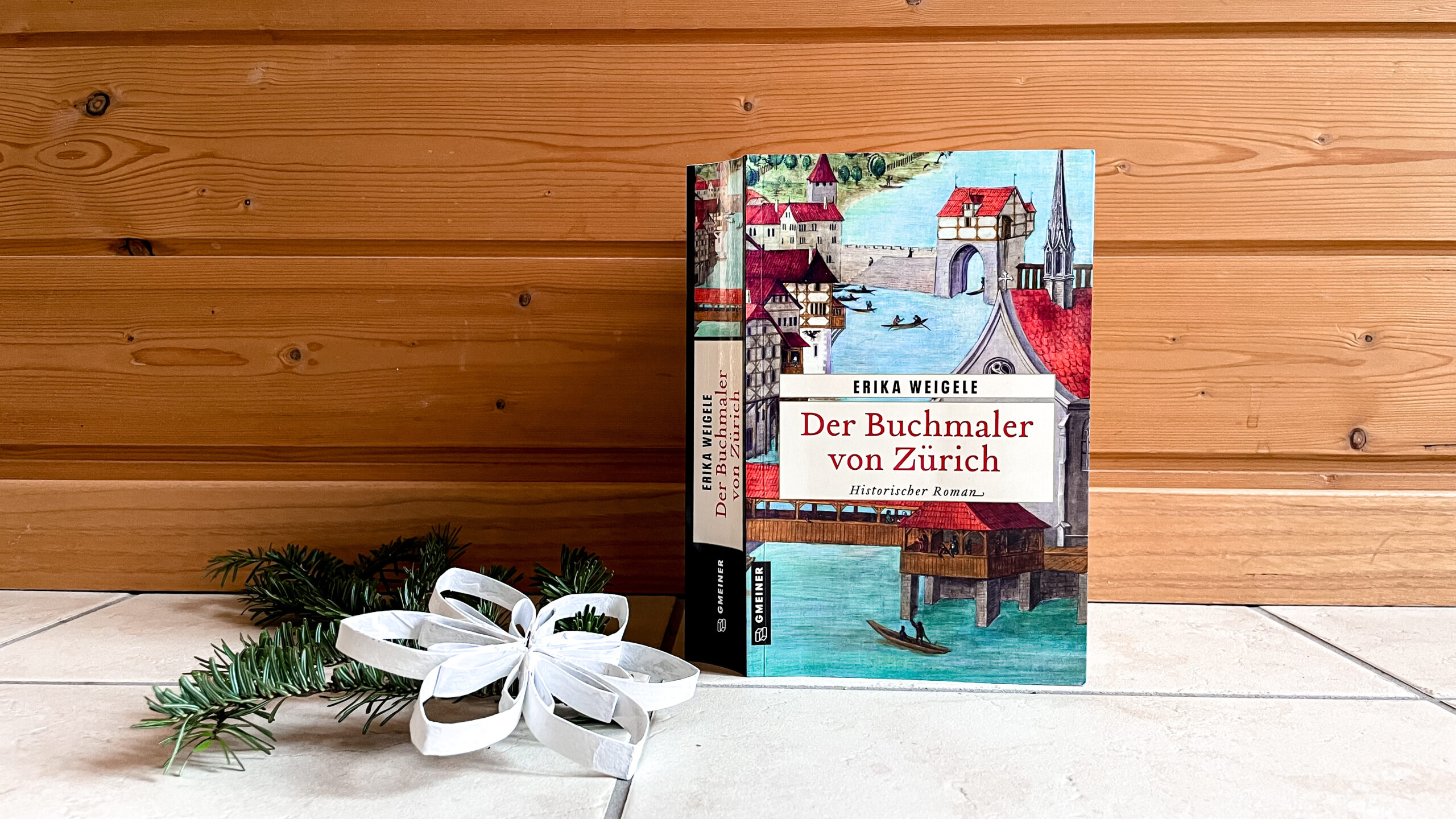 Das Buch «Der Buchmaler von Zürich» steht auf weissen Fliesen an einen Holzwand gelehnt. Daneben liebt ein Tannenzweig und eine aus Karton gebastelte weisse Schneeflocke.