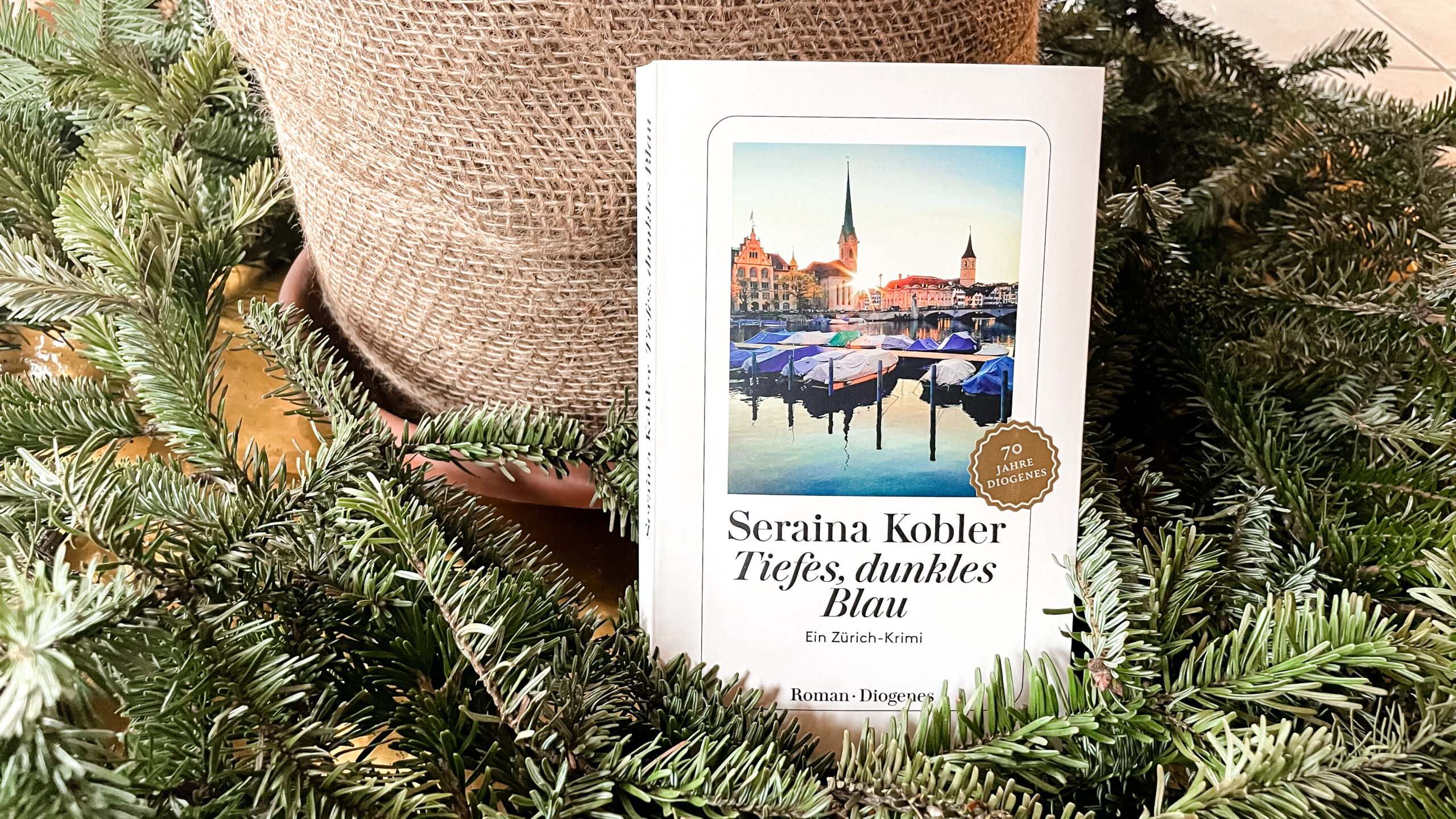 Das Buch «Tiefes, dunkles Blau» von Seraina Kobler steht an einen Pflanzentopf gelehnt inmitten von dunkelgrünen Tannenzweigen.